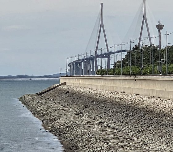 Foto: Straßenverlauf am Meer, Abhang mit Steinen, Brücke und Bäume im Hintergrund sichtbar