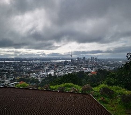 Aufnahme an bewölktem Tag, Blick auf Auckland aus begrünter Umgebung, die Sonne scheint etwas und lässt die Stadt leuchten