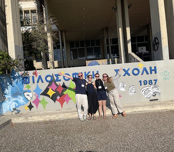 Vortragsgruppe, lächelnd mit teils ausgestreckten Armen, stehen vor einer bemalten Wand welche in griechischer Schrift den Namen der Universität zeigt.